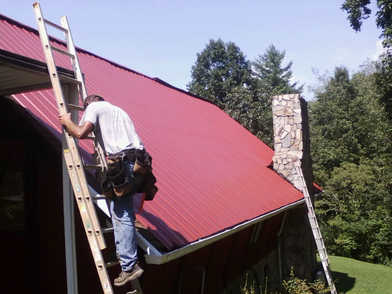 Roofer on a ladder beside a red metal roof and stone chimney — Soffit & Fascia Repair by Roofing Brothers in Piney Point
